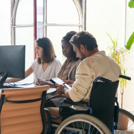 Three diverse individuals, one in a wheelchair, collaborate around a computer and tablet in a modern office, emphasizing inclusion and accessibility.