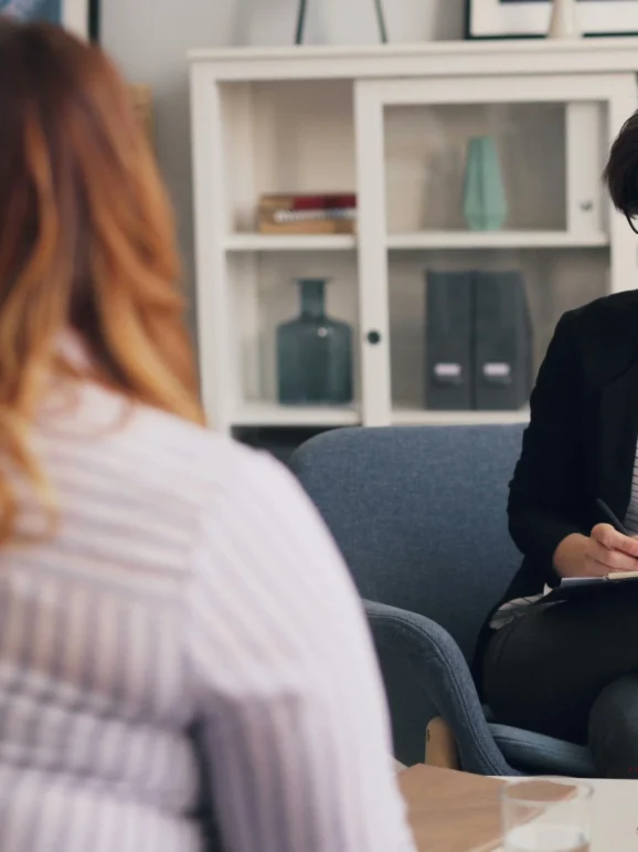 A female therapist with short hair and glasses sits in an office, taking notes and attentively listening to a client seen from the back.