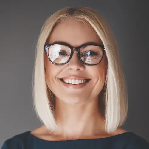A friendly blonde woman with glasses smiles warmly at the camera.