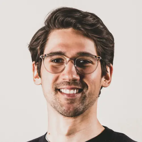 A young man with dark hair and clear-framed glasses smiles warmly.