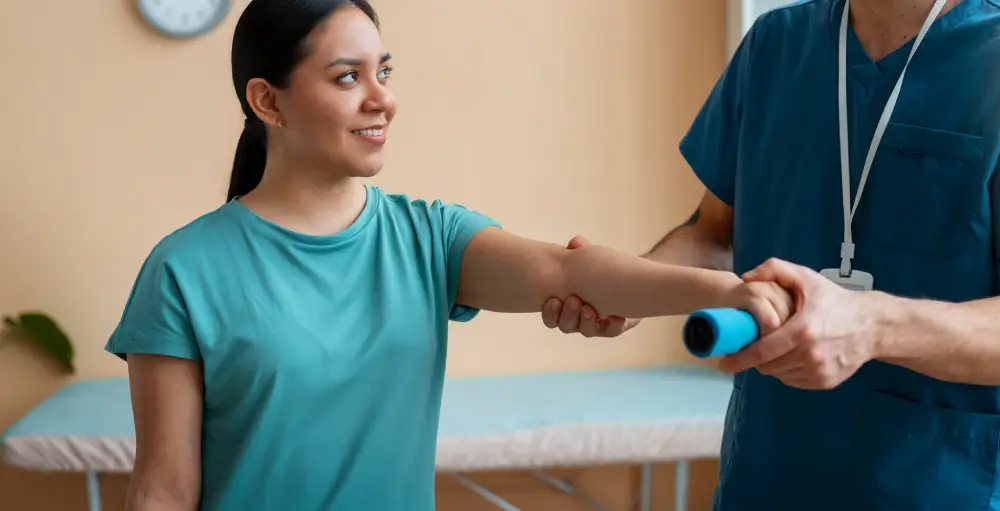 A smiling female patient works on arm rehabilitation with a physical therapist, holding a blue resistance tool.
