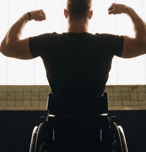Rear view of a man in a wheelchair flexing his muscles in front of a window.