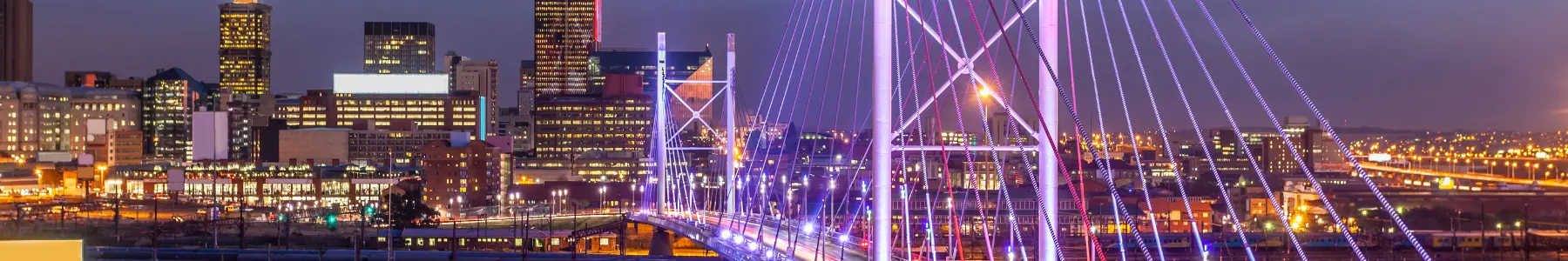 A vibrant night panoramic view of the Nelson Mandela Bridge in Johannesburg, South Africa, lit up in purple, with the city skyline in the background.