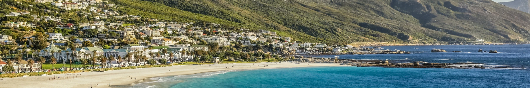 A panoramic view of Clifton Beach in Cape Town, South Africa, featuring a white sand beach, turquoise ocean, luxury homes cascading down a green mountainside, and rocky outcrops.
