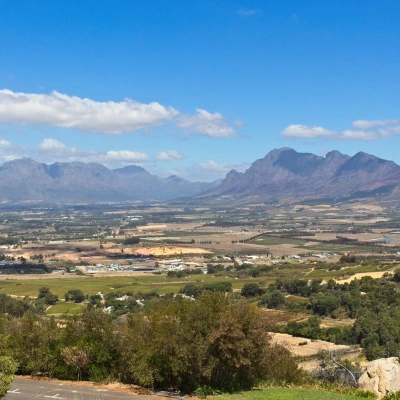 A wide landscape view of a South African valley with vineyards and scattered buildings, framed by majestic mountains under a blue sky with scattered clouds.