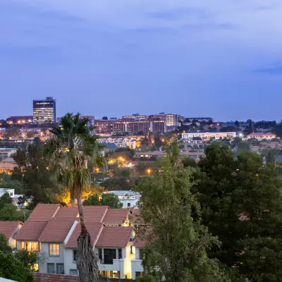 A dusk view of a South African town with red-roofed houses, palm trees, and a lit-up skyline in the distance.