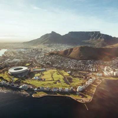 An aerial panorama of Cape Town, South Africa, featuring Table Mountain, the Cape Town Stadium, and the waterfront under clear skies.
