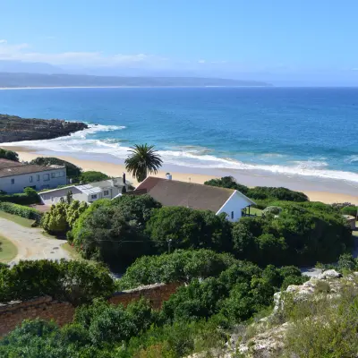 A scenic coastal view with a sandy beach, turquoise ocean, rocky shoreline, and houses nestled among green vegetation.