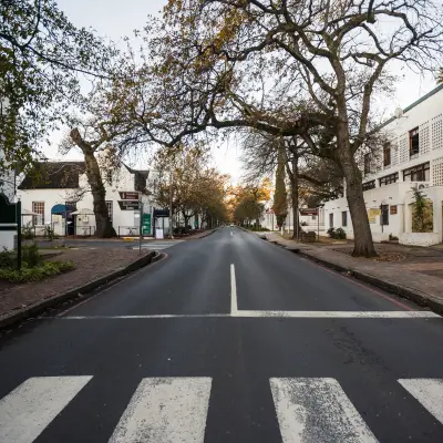 A quiet tree-lined street in a historic South African town, featuring white Cape Dutch style buildings and a pedestrian crossing.