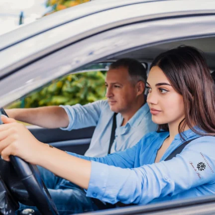 A young woman with dark hair driving a car, looking forward, with a male driving instructor in the passenger seat.