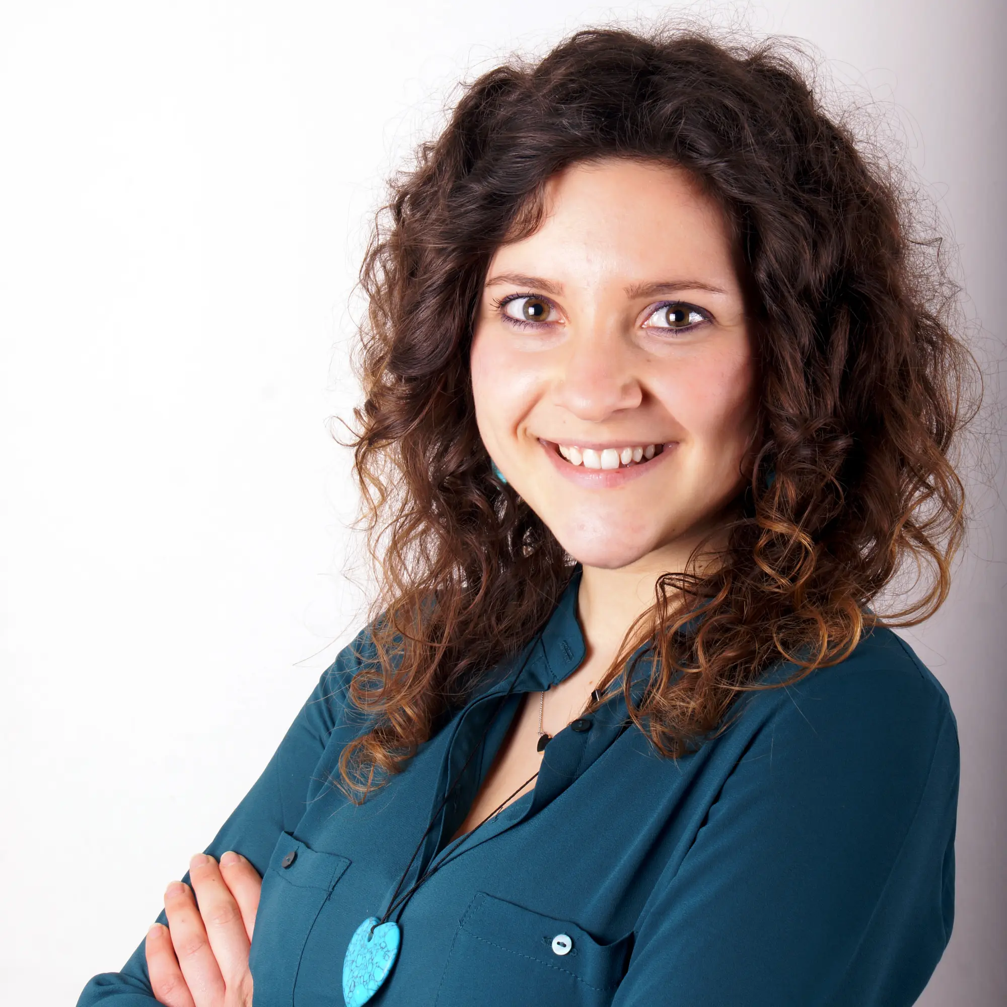 A young woman with curly brown hair and a teal shirt smiles confidently with her arms crossed.