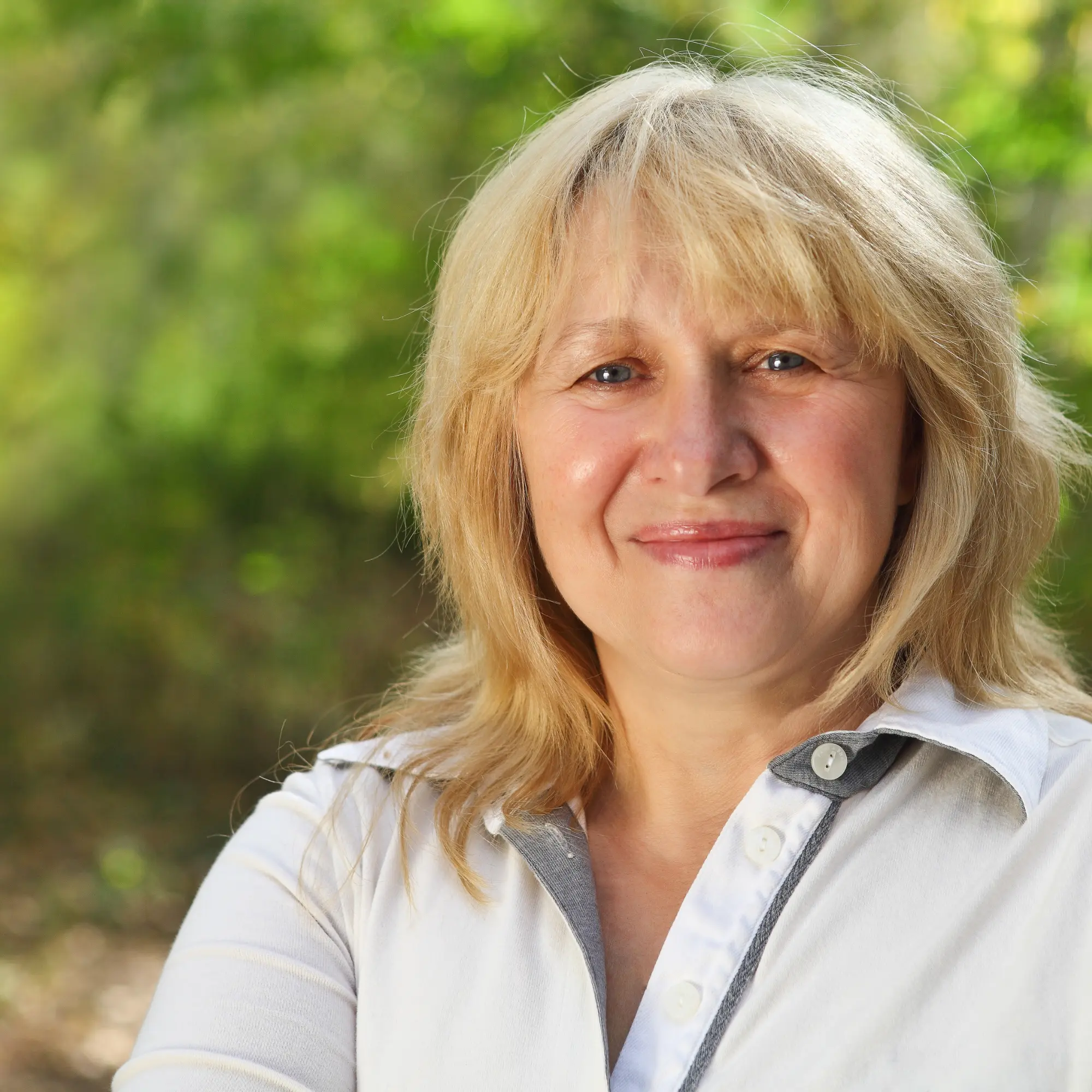 A friendly, smiling blonde woman with blue eyes, wearing a white collared shirt, against a soft green background.